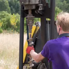 Pracownik w czerwonej rękawicy przygotowuje wiertło do badania geotechnicznego gruntu. Cena takiej usługi zależy od wielu czynników.