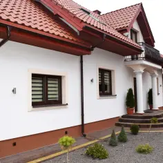 Nowy dom z czerwonym dachem i białymi ścianami, ozdobiony zielenią. Wokół domu ścieżki z kostki brukowej i małe drzewka.