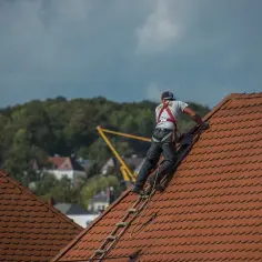 Dekarz w uprzęży bezpieczeństwa pracuje na czerwonym, dachówkowym dachu.