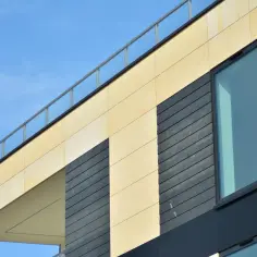 Nowoczesna elewacja wentylowana z jasnymi panelami i ciemnymi elementami, dużymi oknami i szklaną balustradą na tle błękitnego nieba.
