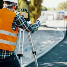 Geodeta w kamizelce odblaskowej z niwelatorem na statywie, pokazuje kciuk w górę.