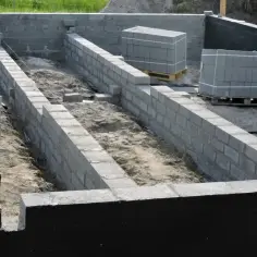 Budowa domu w toku: wykopy pod fundamenty i pierwsze ściany z bloczków betonowych. Materiały budowlane czekają na użycie.