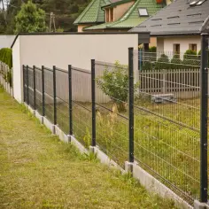 Betonowe bloczki jako co zamiast podmurówki pod ogrodzenie. Nowoczesne ogrodzenie z siatki na tle domów.