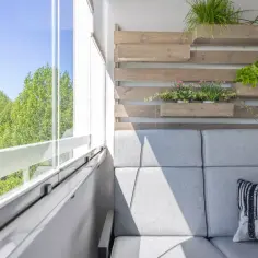 Przytulna loggia z szarą sofą, poduszką z liskiem i zielenią na drewnianej ściance. Widok na drzewa.