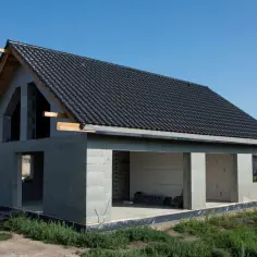 Dom w stanie surowym otwartym, z betonowych pustaków, z czarnym dachem z dachówki. Widać otwory okienne i drzwiowe.