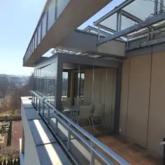 Nowoczesny ogród zimowy z przeszklonymi ścianami, stołem i krzesłami, oferujący widok na osiedle i plac zabaw.