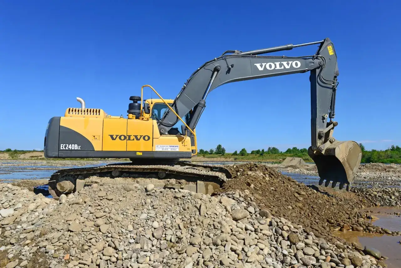 Żółta koparka Volvo EC240B LC pracuje przy rzece, przesypując kamienie i ziemię.