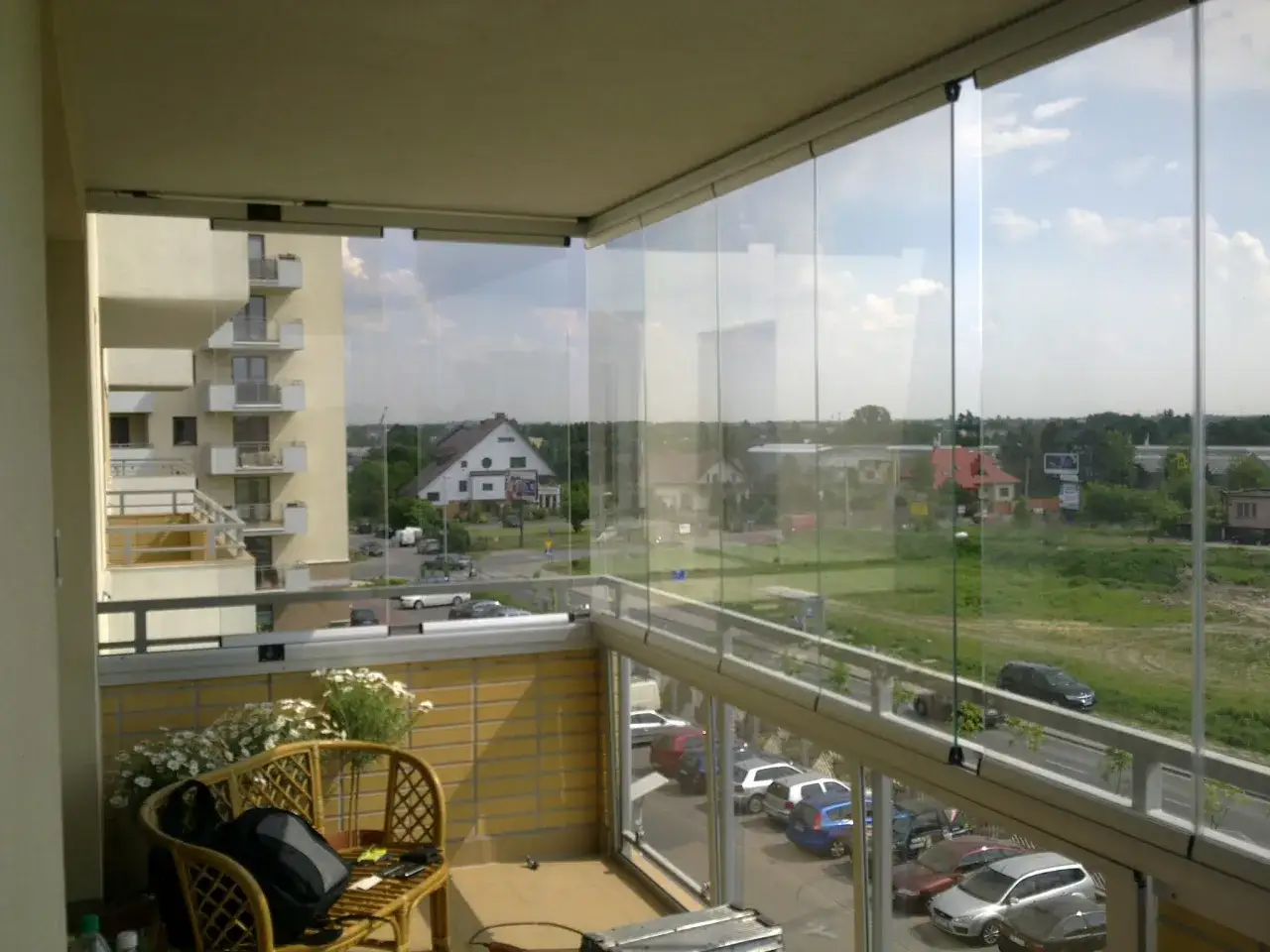 Zabudowa balkonu w bloku z widokiem na ulicę i inne budynki. Na balkonie rattanowe krzesło z plecakiem i kwiatami.