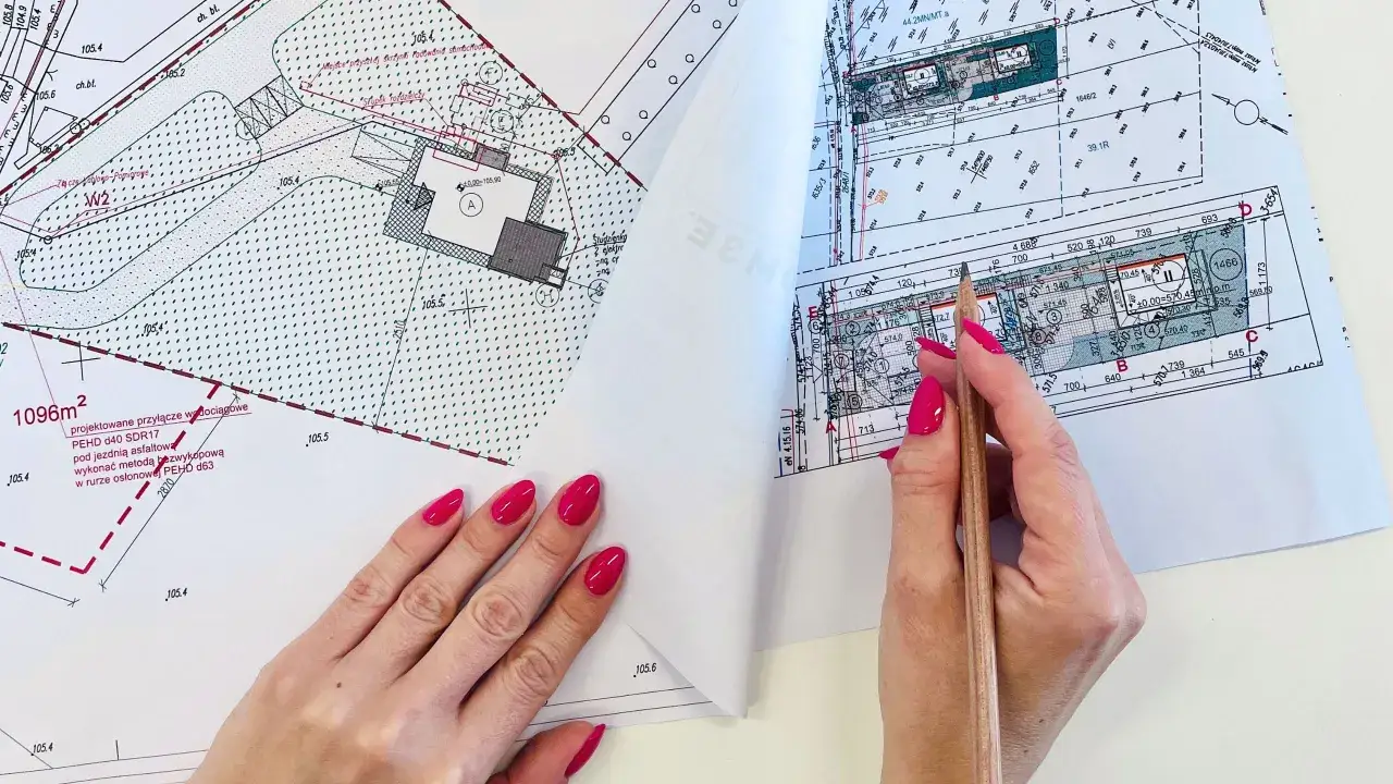 Ręce z różowym lakierem przeglądają mapę do celów projektowych, wskazując ołówkiem szczegóły planowanej inwestycji.
