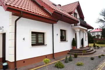 Nowy dom z czerwonym dachem i białymi ścianami, ozdobiony zielenią. Wokół domu ścieżki z kostki brukowej i małe drzewka.