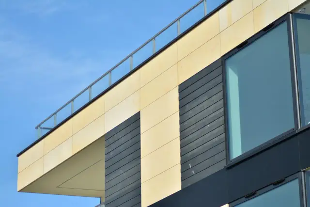 Nowoczesna elewacja wentylowana z jasnymi panelami i ciemnymi elementami, dużymi oknami i szklaną balustradą na tle błękitnego nieba.